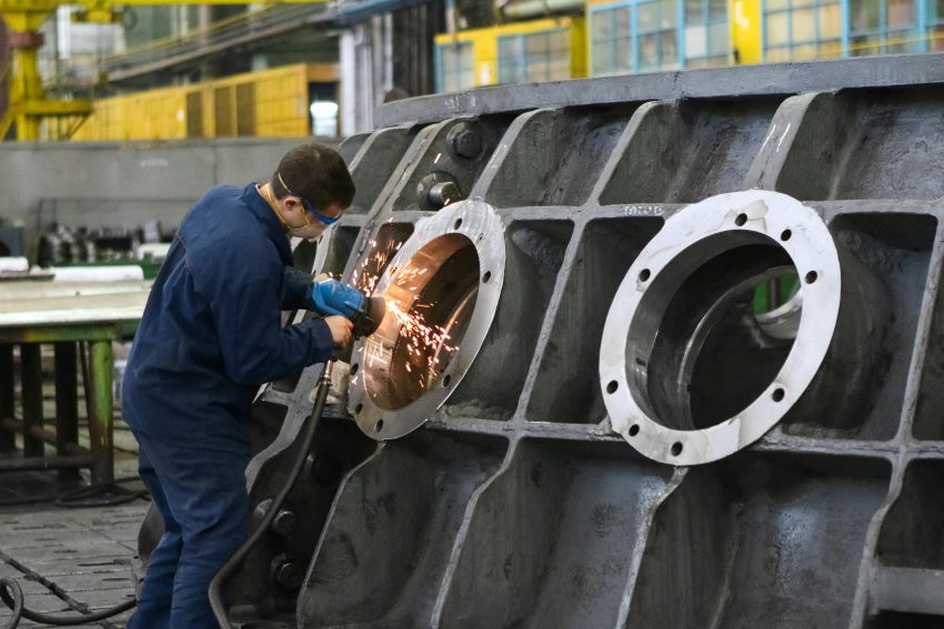 Wielki metalowy kadłub drążony i szlifowany przez pracownika w kombinezonie zajmującego się produkcja wyrobów metalowych Toruń