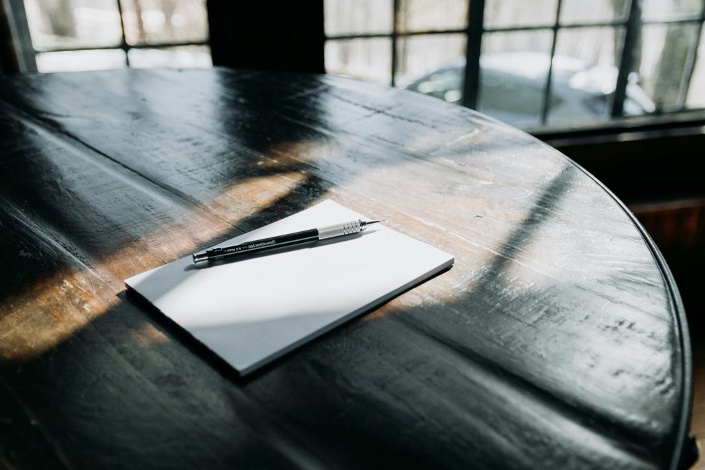 Na drewnianym stole leży biała kartka papieru, na której położony jest długopis