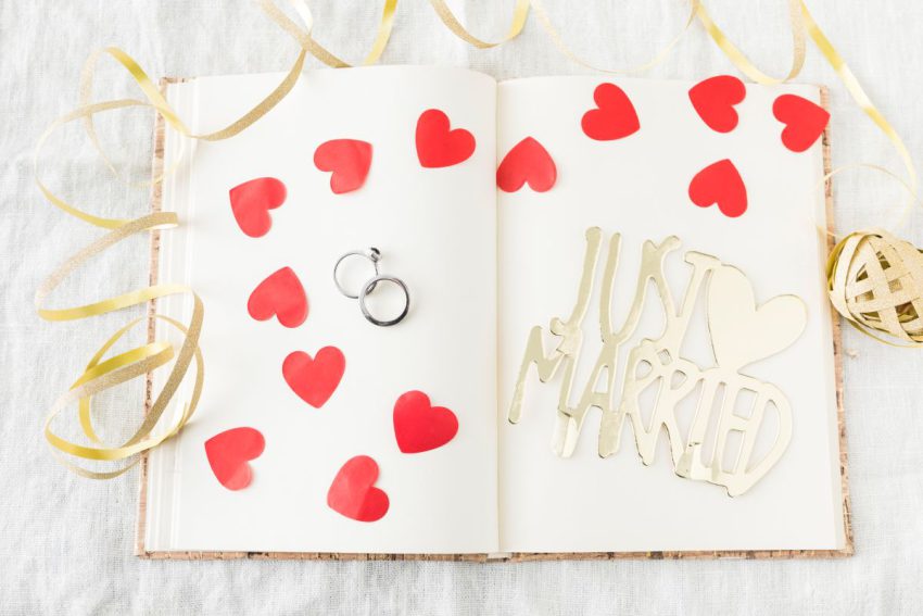 Otwarta księga gości z białymi kartami, na których rozsypano czerwone papierowe serca. Obok znajdują się srebrne obrączki oraz złoty napis dekoracyjny "Just Married".